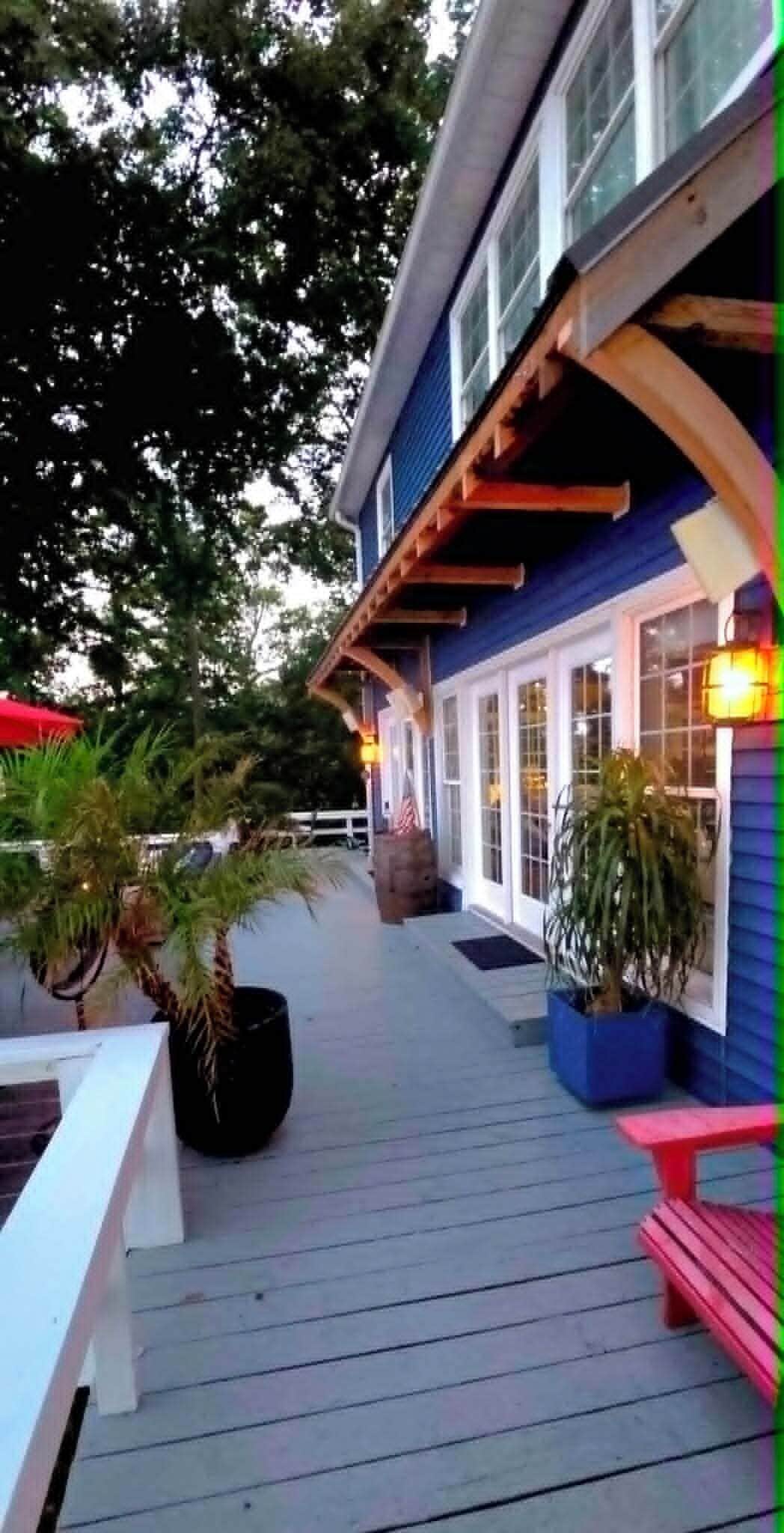 A wooden deck with potted plants and a bench, leading to a building with a blue exterior and large windows.