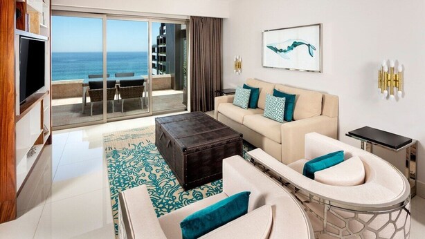 Ein modernes Wohnzimmer mit einer Couch, einem Couchtisch und einem Fernseher. Durch große Fenster ist der Blick aufs Meer möglich.