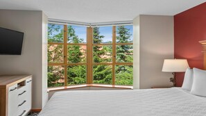 Ein Hotelzimmer mit großem Erkerfenster, einem Bett, einem Nachttisch, einer Lampe und einem Flachbildfernseher.