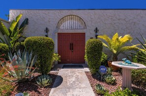 A building with a red door, a stone wall, and various plants. - Stylish Spanish townhome w/ mountain views, pool & spa in South Palm Springs (Palm Springs)