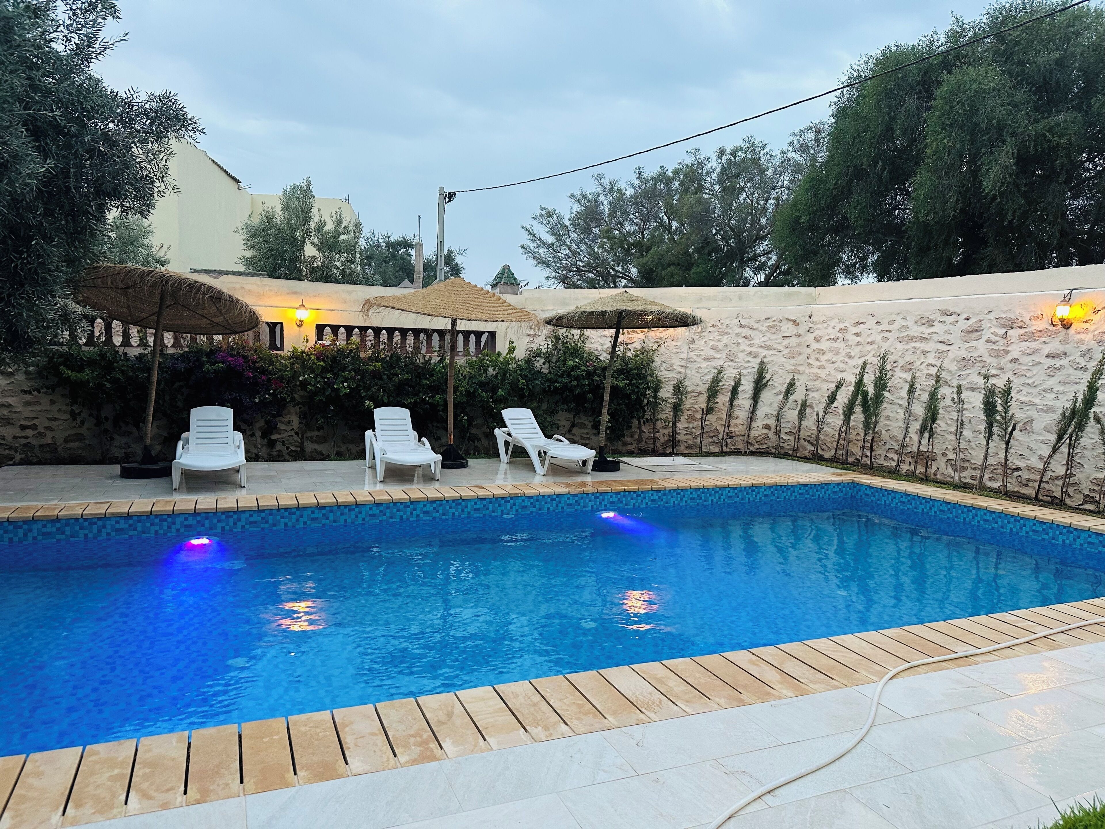 Piscina con sillas de descanso y sombrillas, rodeada de un muro de piedra y vegetación.