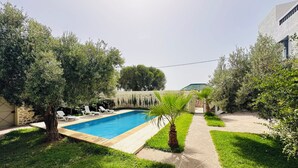 A rectangular swimming pool with a wooden deck, surrounded by well-maintained grass and trees. - Villa Targante, Essaouira (Essaouira)