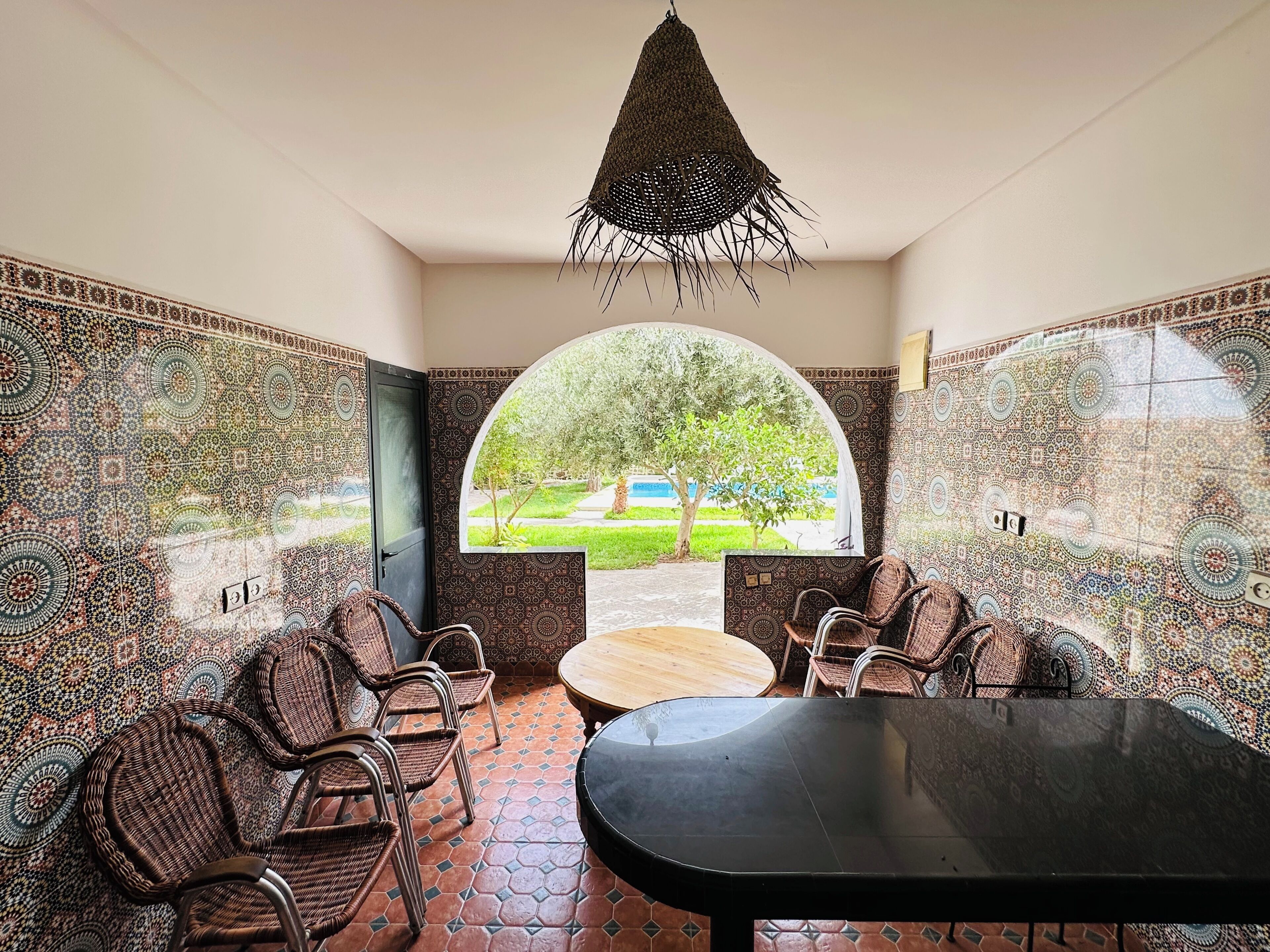 A room with patterned walls, a round table, wicker chairs, and a view through a semi-circular window.