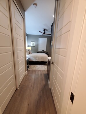 Um corredor estreito que dá para um quarto, com cama, ventoinha no teto e janela com persianas.