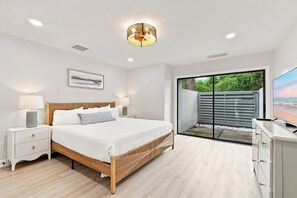 Ein Schlafzimmer mit einem Bett, Nachttischen, einer Lampe, einem Gemälde, einem Fernseher und einer Schiebetür zu einem Außenbereich.