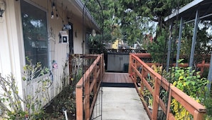 Un sendero de hormigón que lleva a una terraza de madera con barandilla metálica, rodeada de follaje y un edificio con ventana y puerta.