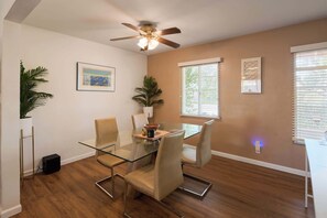 A dining area with a glass table, cushioned chairs, a ceiling fan, and a window with blinds. - Backdrop - Private Pool - Mins to Old town (Scottsdale)