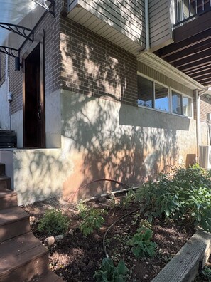 A residential building with a brick facade, a wooden front porch, and a garden area with plants and a hose. - Renovated and private garden apartment near UofU. (Salt Lake City)