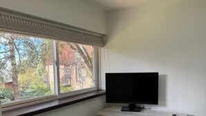 Ett rum med ett fönster, ett element, en vit stol, ett skrivbord med en tv och en fjärrkontroll.