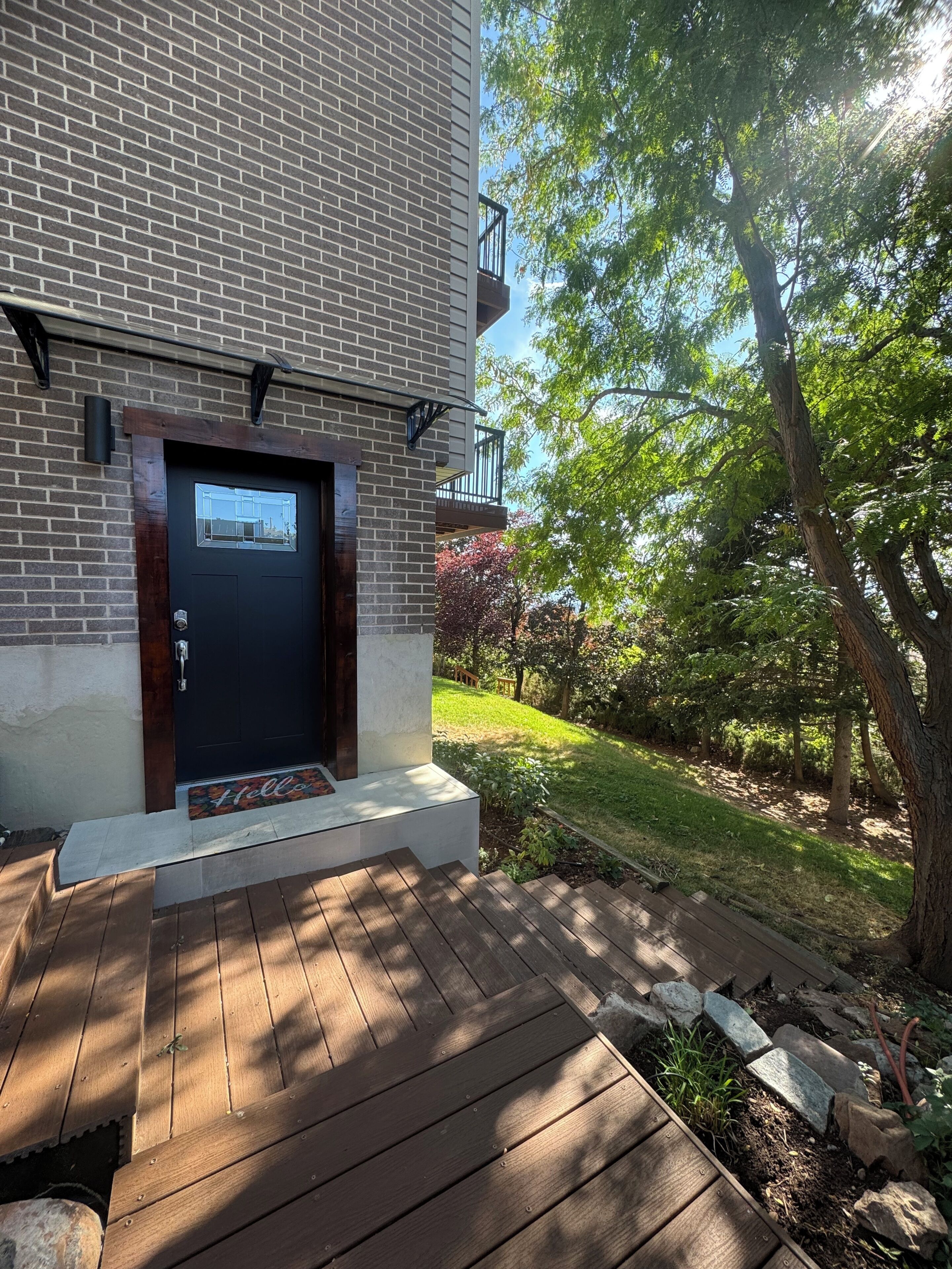 A modern brick building with a dark front door, a small porch with a welcome mat, and a well-maintained garden with trees and a lawn.