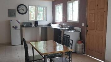 Een kleine keuken met een koelkast, magnetron, fornuis en spoelbak. Er is een eettafel met stoelen.