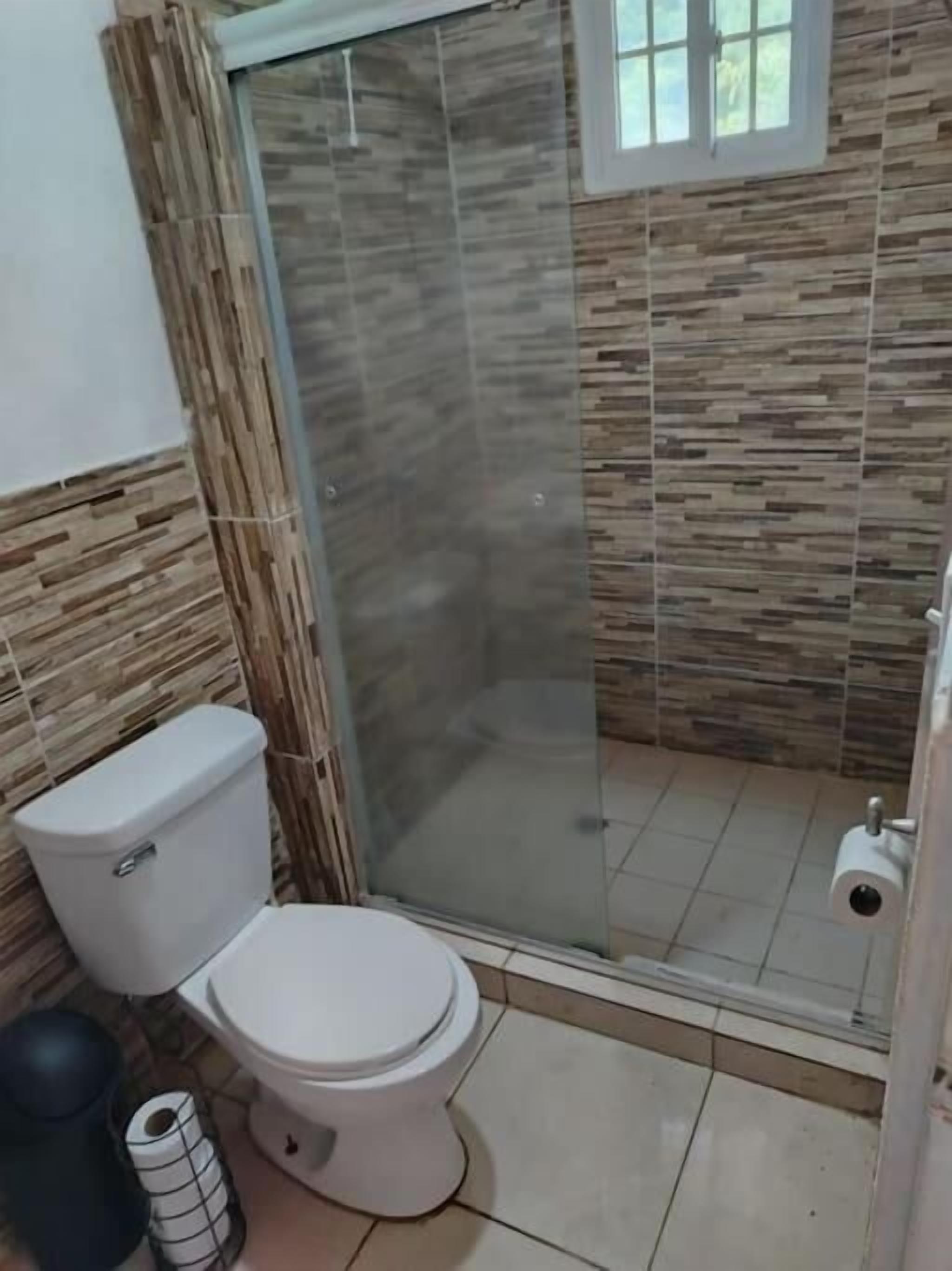 Baño con inodoro, cabina de ducha de vidrio y dispensador de papel higiénico.