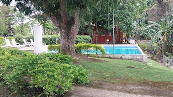 Zona de piscina con un edificio rojo al fondo, rodeada de exuberante vegetación y una estructura blanca.