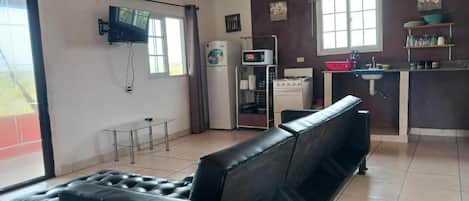 A living room with a black leather sofa, a television mounted on the wall, a kitchen area with a sink and cabinets, and a dining table.