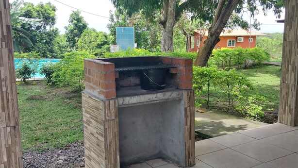Una parrilla de ladrillo para exterior con rejilla metálica, rodeada por un jardín con diversas plantas y un patio con baldosas.