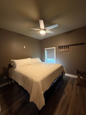 Un dormitorio con cama, ventilador de techo, ventana y perchero con ganchos.