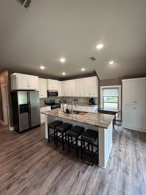 A modern kitchen with white cabinets, a stainless steel refrigerator, a granite countertop island with bar stools, and a window providing a view. - Blue House on the Corner in Old Higden Perfect 4-bedroom house with AC (Higden)