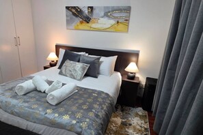 Habitación de hotel con cama, dos mesitas de noche con lámparas, un cuadro sobre la cama y una cortina a la derecha.