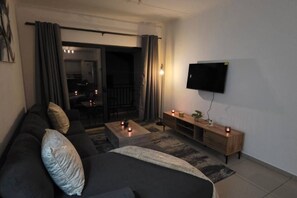 A living room with a sofa, coffee table, TV, and a balcony with curtains. - Stylish 2BR Apartment, Pool & Balcony, Joburg (Midrand)