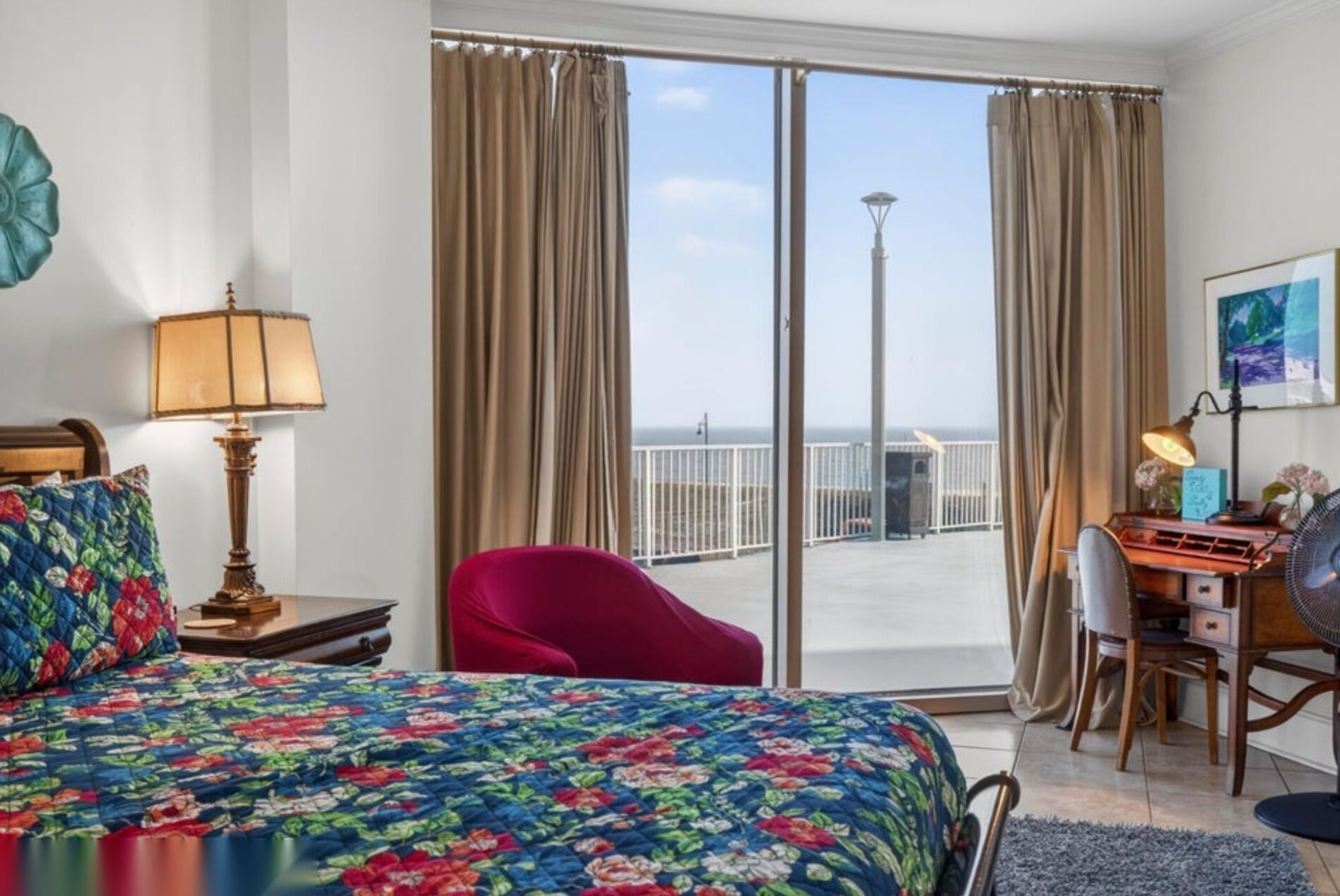 A hotel room with a bed, a desk, a chair, and a view of the sea through large windows.