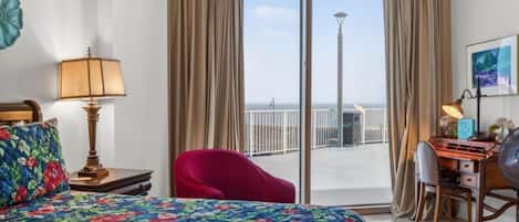A hotel room with a bed, a desk, a chair, and a view of the sea through large windows.