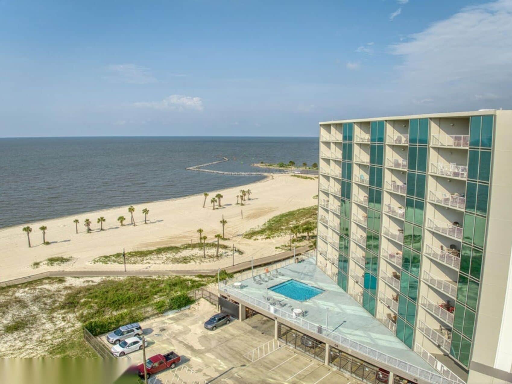 A hotel with a pool on the ground floor, a beach view, and a parking area with cars.