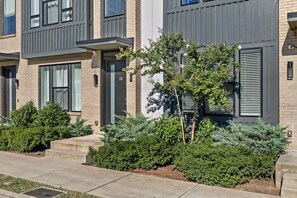 A modern residential building with a grey facade, a visible entrance, and a well-maintained garden with various shrubs and a small tree. - City View Rooftop Retreat w/ FirePit • Close to DT (Nashville)