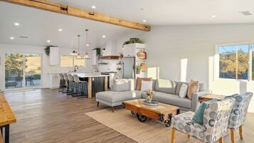 Un amplio salón con sofá, mesa de centro y sillones, una cocina con armarios blancos y un ventanal que proporciona luz natural.