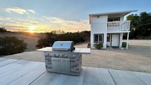 An outdoor cooking area with a grill and a built-in oven, a two-story white house with a balcony, and a sunset in the background. - The Shire at Baggins Hill by Avantstay Hot Tub + Playhouse + Cornhole 10 Private Acres (Templeton)