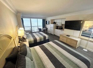 A hotel room with two beds, a desk, a dresser, a TV, and a balcony with a view. - Surf N Sun spectacular oceanfront studio w/balcony (Daytona Beach)