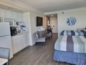 Une pièce compacte comprenant un coin cuisine, un lit, une chaise et un téléviseur fixé au mur.