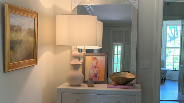 Een hal met een witte dressoir, een lamp, een ingelijst beeld en een potplant.