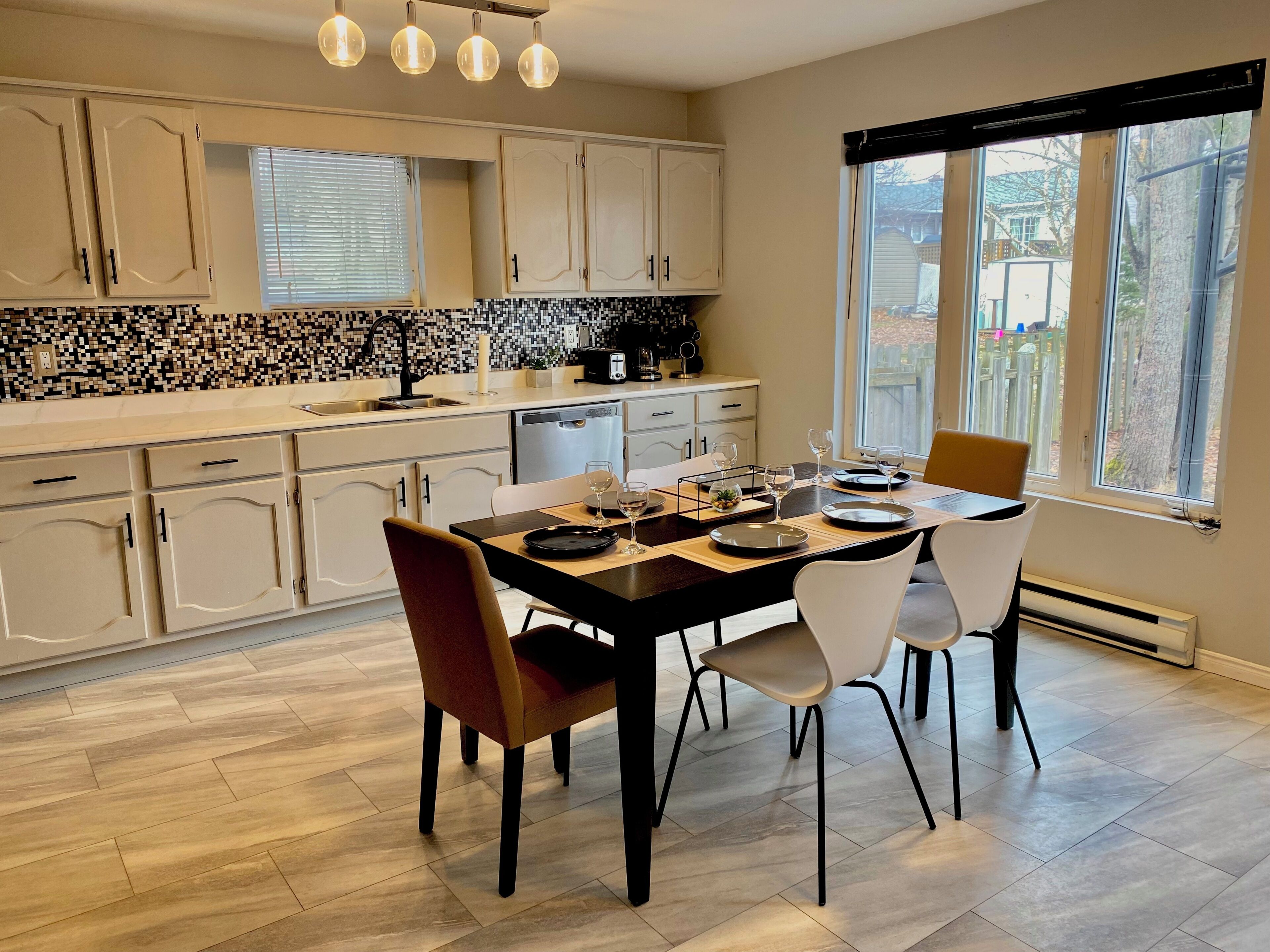 A modern kitchen with a dining area, featuring a tiled backsplash, wooden cabinets, and a large window with a view.
