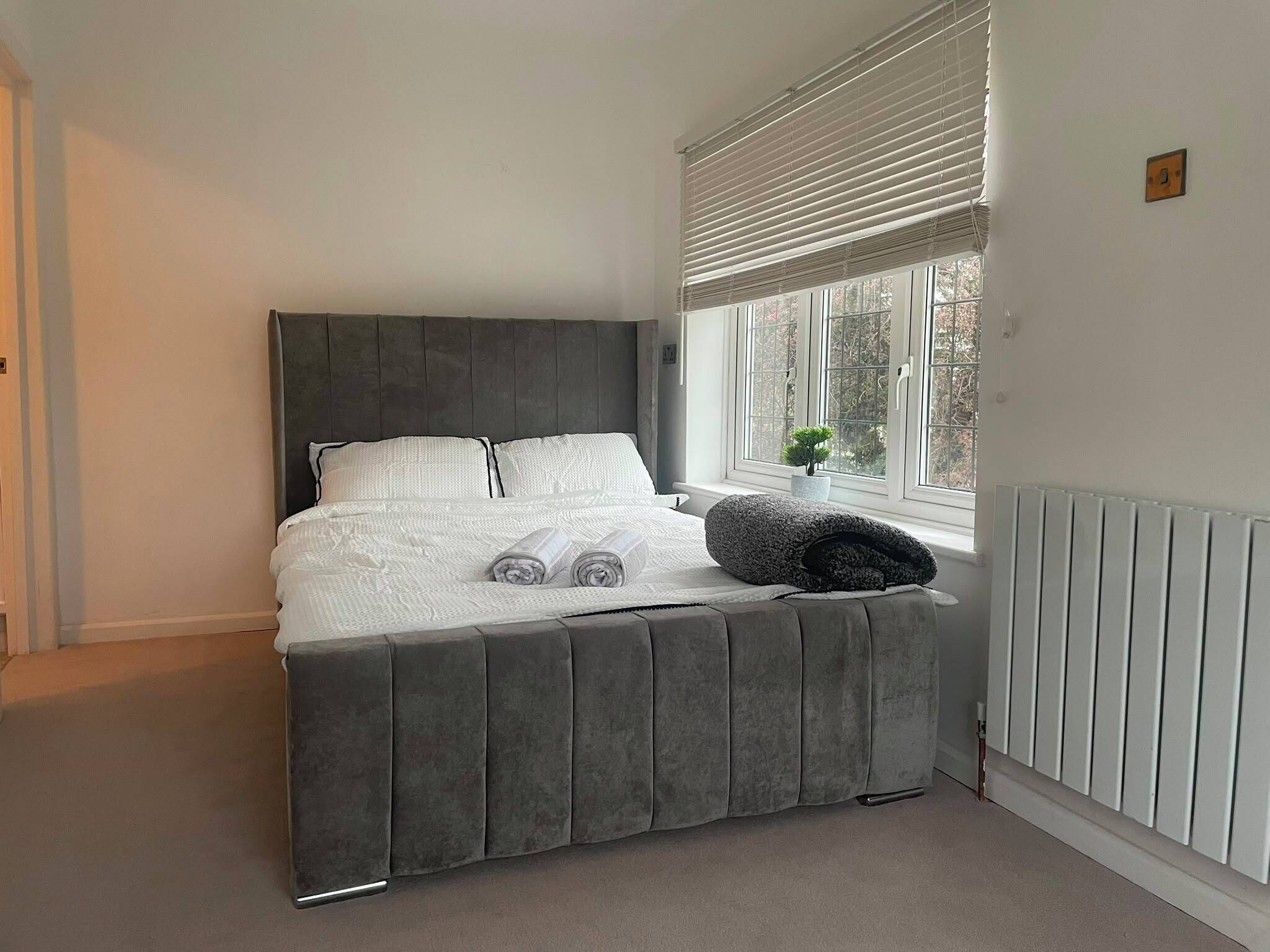 A bedroom with a bed, a window with blinds, a radiator, and a small plant on the windowsill.