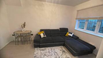 A modern living room with a sectional sofa, a dining table with chairs, and a large window with blinds.