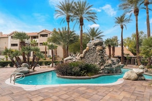 A swimming pool with a rock waterfall feature, surrounded by palm trees and a building with a tiled roof. - Luxury 2BR/2BA Condo - Walk to the Strip, Rio & Palms! (Las Vegas)
