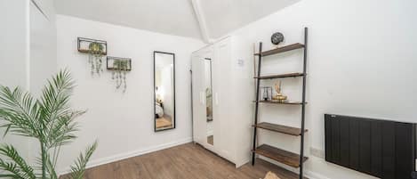A hallway with wooden flooring, white walls, and a black radiator. There are two wall-mounted shelves with decorative items and a mirror reflecting a room.