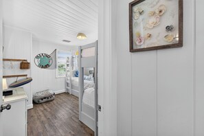 A room with a bunk bed, a wall-mounted shelf, a framed artwork, and a door leading to another room. - Sugar Magnolia! Beach Front, Private Pool, Free Beach Gear! (St. George Island)