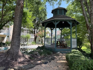 A gazebo with a wooden bench and a small table, surrounded by trees and a garden.