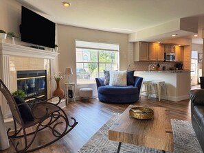 A living room with a fireplace, a television mounted on the wall, a blue armchair, and a kitchen area with a microwave and sink.