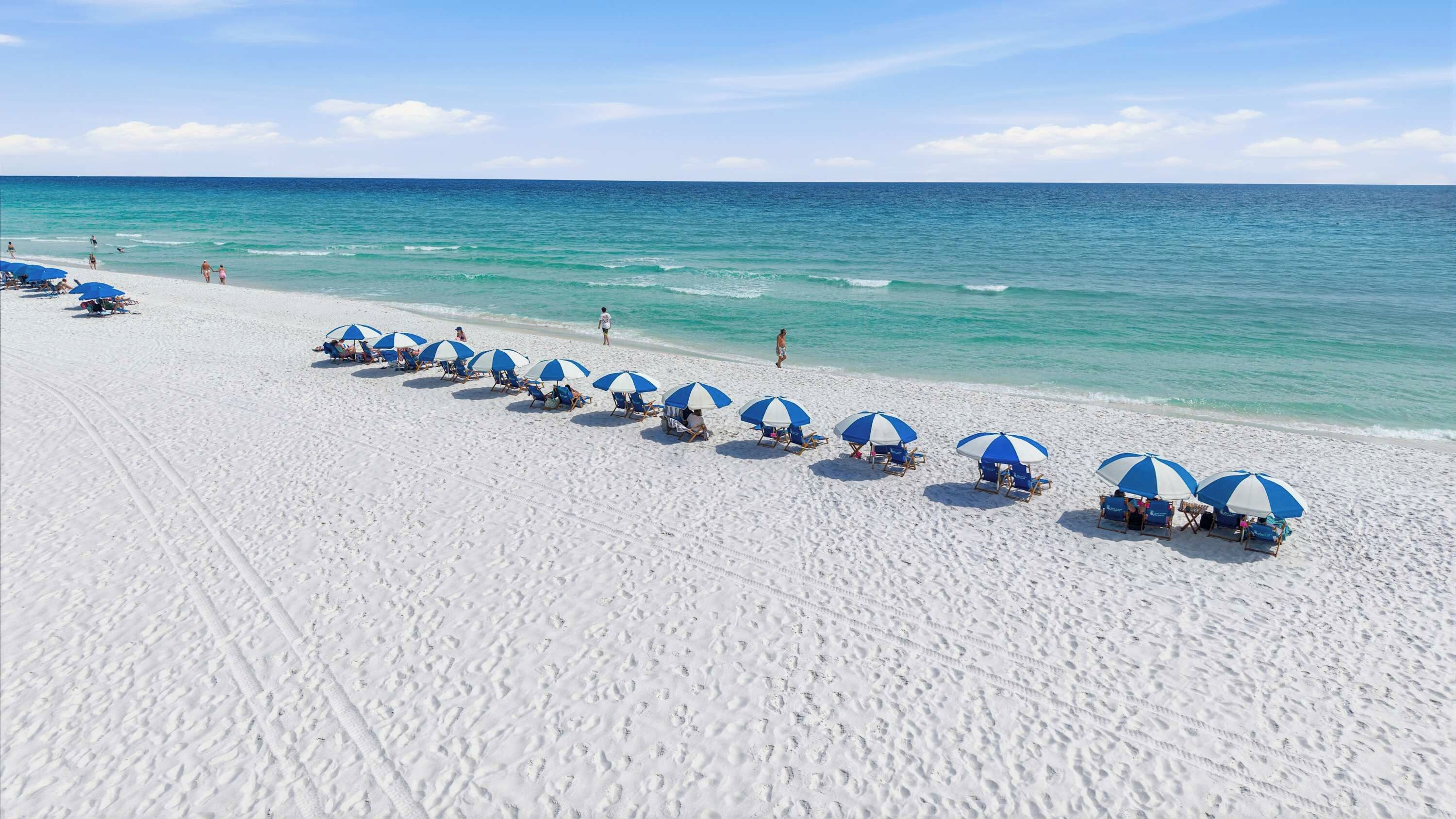 Ein Strand mit blauen und weißen Sonnenschirmen, weißem Sand und dem Meer.