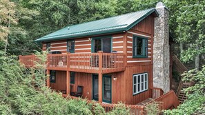 A two-story cabin with a balcony, surrounded by dense forest.
