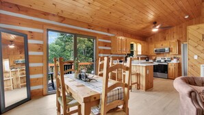 A wooden cabin interior with a dining area, kitchen, and large sliding glass doors leading to an outdoor deck. - 3BR mountain-View cabin with hot tub, fireplace, WiFi, and Views (West Jefferson)