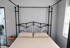 Una cama individual con estructura metálica y ropa de cama color beige en una habitación con pared lisa y una ventana.