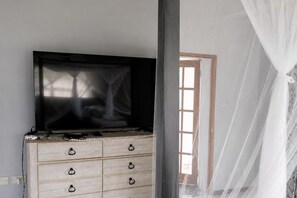 Ein Zimmer mit Fernseher auf einer Holzkommode, einem Fenster mit durchsichtigen Vorhängen und Blick auf ein Gebäude.