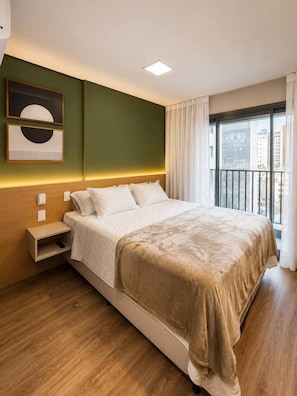 Ein Hotelzimmer mit Bett, Nachttisch, Wandbild und einem Fenster mit Vorhängen.