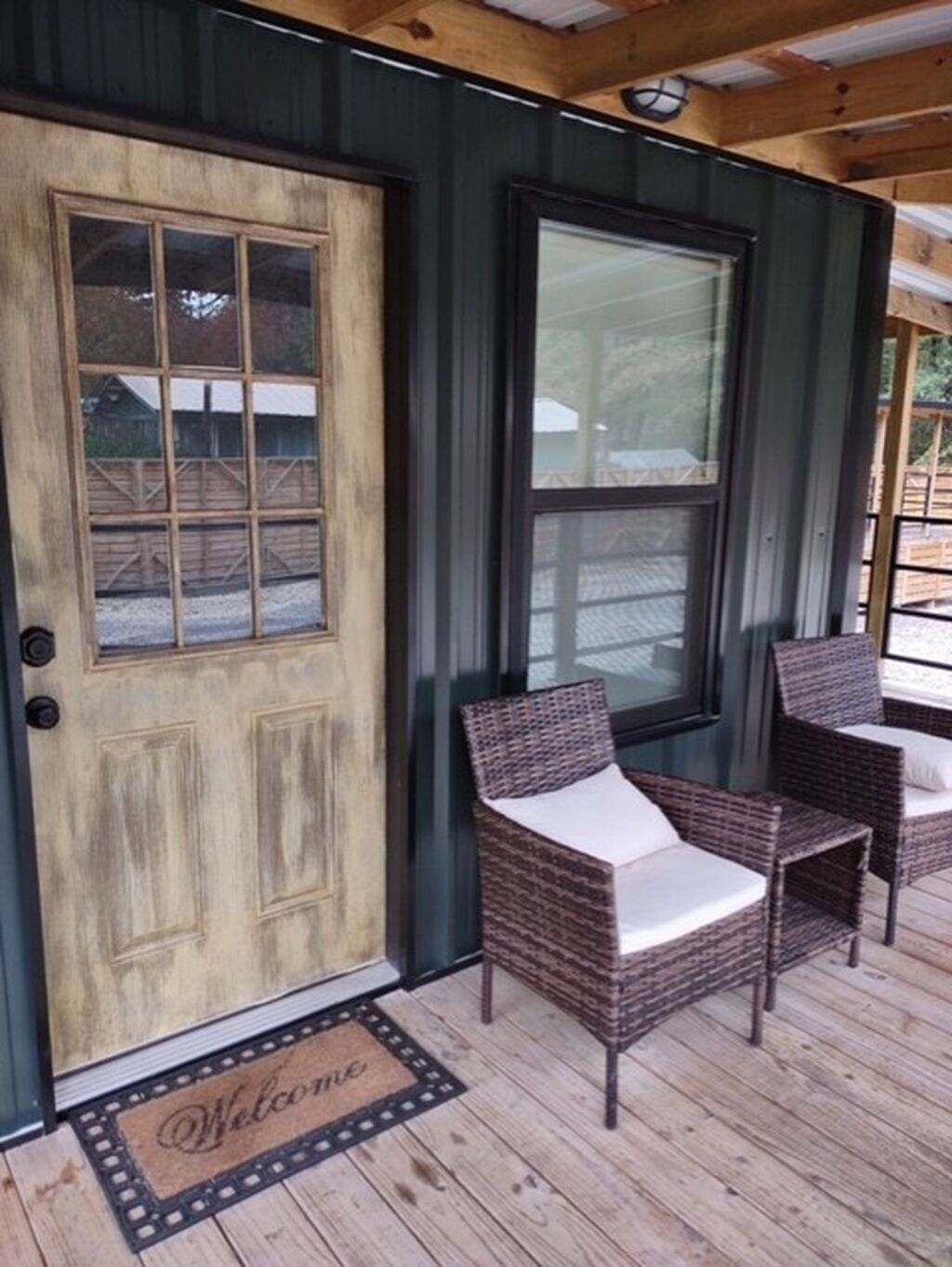 A wooden door with a window, two wicker chairs with cushions, and a welcome mat on a wooden deck.