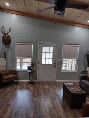 A room with a ceiling fan, a deer head decoration, two windows with blinds, a door, a brown leather chair, a small table with a plant, and a wooden bench.