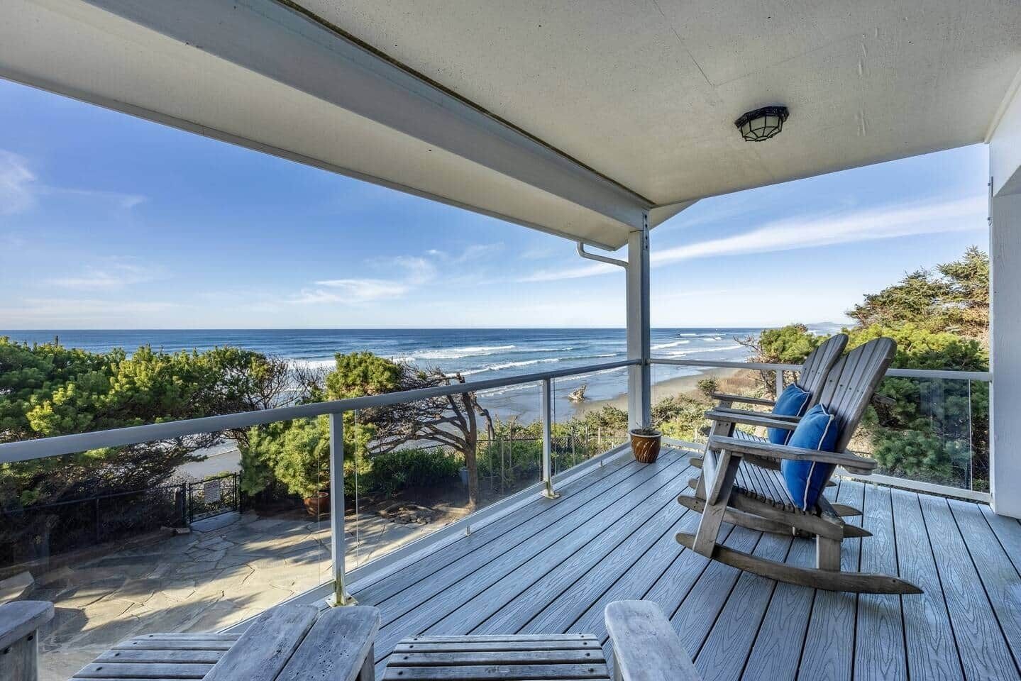Une terrasse en bois avec un fauteuil à bascule et un pot de fleurs, donnant sur une plage avec vue sur l’océan.