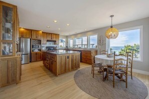 A modern kitchen with wooden cabinets, a central island, and a dining area with a view of the ocean. - Sanctuary at Seal Rock (Seal Rock)